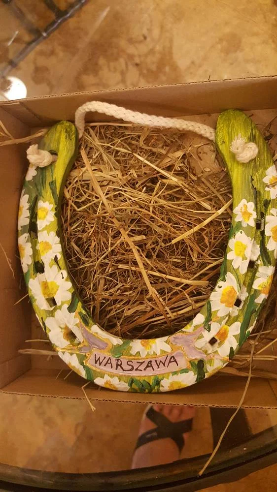 Podkowa z białymi kwiatami, zielonymi liśćmi i napisem „Warszawa”, ułożona w sianie.