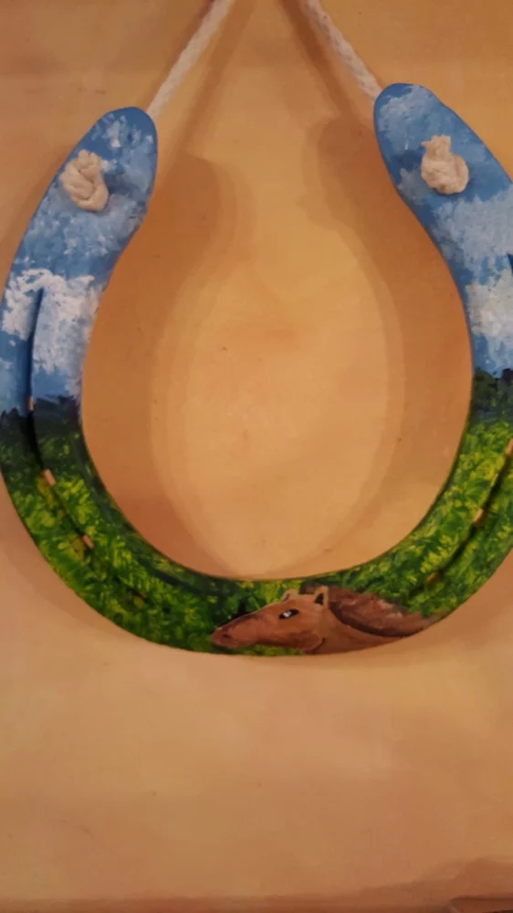 Podkowa z błękitnym niebem, zieloną łąką i dwoma końskimi profilami u dołu.